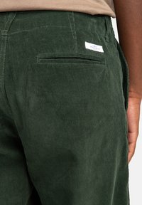 Dark green corduroy pants feature a rear patch pocket with a small white logo tag and a textured ribbed surface.
