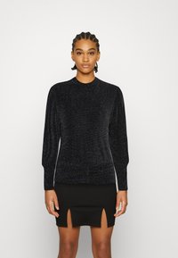 Blusa preta de manga longa com um pescoço alto, feita de um tecido texturizado e brilhante. Combinada com uma saia preta com fendas laterais.