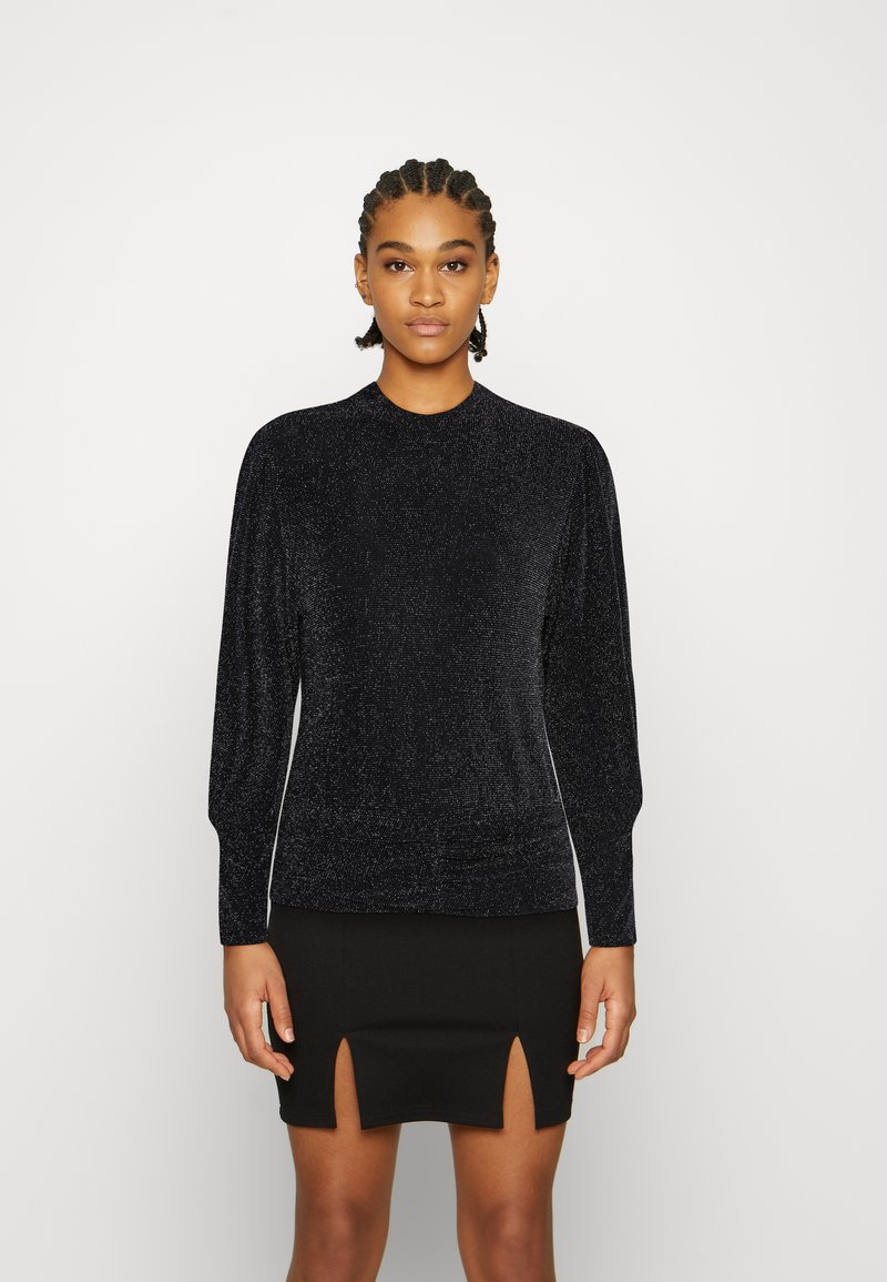 Blusa preta de manga longa com um pescoço alto, feita de um tecido texturizado e brilhante. Combinada com uma saia preta com fendas laterais.