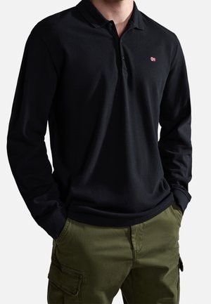 Mand iført sort langærmet polo med lille flaglogo og olivengrønne cargobukser, hænderne i lommerne, stående.