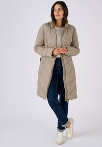 Beige gewatteerde donsjas met een hoge kraag en volledige ritssluiting, gecombineerd met donker gewassen jeans en witte sneakers met beige accenten.