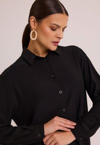 Czarna koszula zapinana na guziki z klasycznym kołnierzykiem, długimi rękawami i czarnymi guzikami. Modelka nosi duże okrągłe kolczyki. Gładka tekstura materiału.