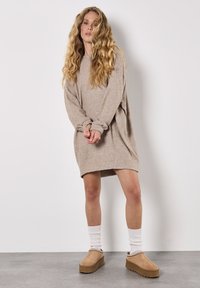 Beige oversized sweatshirt jurk met lange mouwen, een ontspannen pasvorm en geribbelde boorden, gecombineerd met tan platform schoenen en witte sokken.