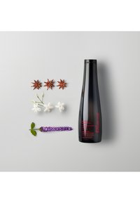 Bottiglia di shampoo nera con testo rosso, circondata da fiori bianchi, lavanda viola e anice stellato, su uno sfondo grigio chiaro.