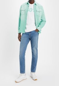 Chemise en denim vert clair portée sur un t-shirt blanc avec texte, associée à un jean bleu et des baskets blanches avec semelle gomme.