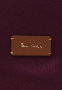 Brown leather label with yellow stitching displaying "Paul Smith" in gold script, placed on a textured maroon fabric background.