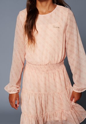 Mujer vestida con un vestido de manga larga color rosa claro con un sutil patrón repetido de logotipos y cintura y puños fruncidos.