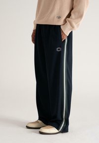Man in een beige gebreid sweater en zwarte wijde broek met groene en witte zijkanten, staand met handen in de zakken en beige schoenen.