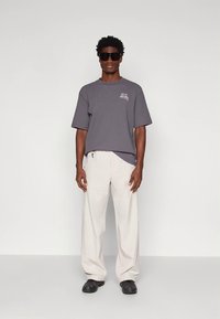 Gray ribbed short-sleeve shirt with white text, paired with loose white trousers and black shoes, worn by a standing model.