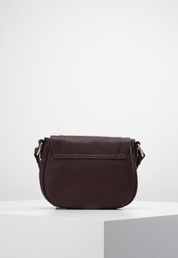 Tmavě hnědá kožená kabelka přes tělo s klopou a zlatými kovovými detaily, vystavená na bílém podstavci na jednolitém pozadí.