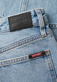 Los jeans de mezclilla de color azul claro cuentan con una parche de cuero negro con texto en relieve y una etiqueta rosa de "SUPERDRY" en el bolsillo delantero.