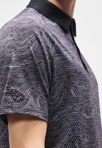 Gros plan d'une personne portant un polo à manches courtes avec un col noir et un motif hexagonal, arborant un logo sur la manche.