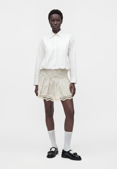 Femme à la peau foncée portant un chemisier blanc à manches longues, une jupe courte rayée avec bordure en dentelle, des chaussettes blanches et des chaussures noires, debout devant un fond uni.