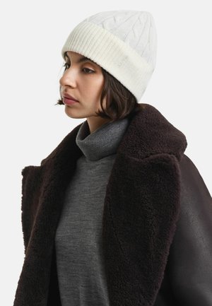 Weiße Strickmütze mit Zopfmuster und einem gerippten Bund. Das Modell trägt einen grauen Rollkragenpullover und einen dunkelbraunen, flauschigen Mantel.