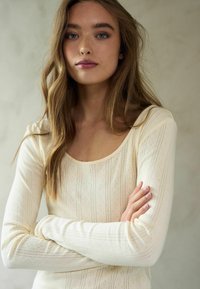 Ilgiem piedurknēm tops krēmkrāsā, ar izteikta izgriezuma kakla daļu, ribotu tekstūru un smalkām vertikālām svītrām ar delikātiem rakstiem.