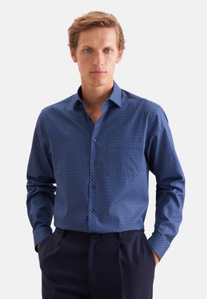 Homme portant une chemise bleue à motifs à manches longues, boutonnée et rentrée dans un pantalon bleu marine foncé, les mains dans les poches.