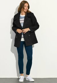 Abrigo de lana negro tipo peacoat con cuello ancho y botones dobles, llevado sobre un suéter estampado. Combinado con pantalones slim azul oscuro y zapatillas blancas.