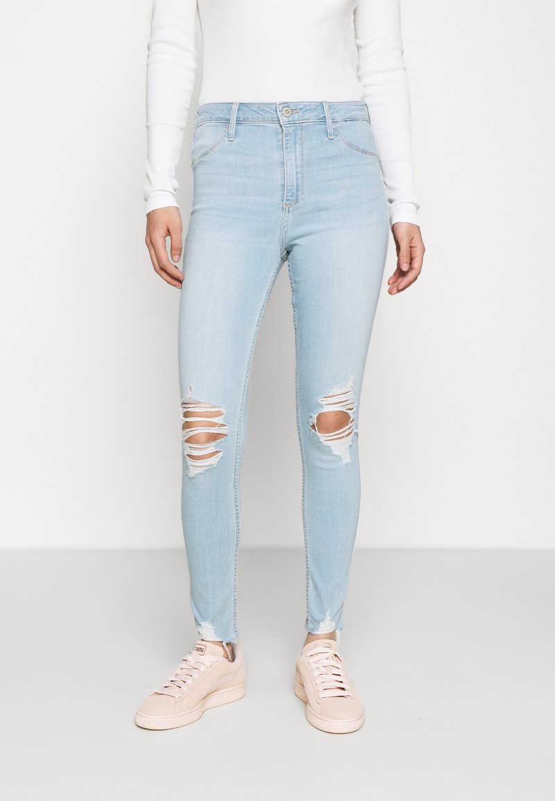 Mujer llevando jeans skinny rasgados de color azul claro, blusa blanca de manga larga y zapatillas Puma rosas claras contra un fondo simple.