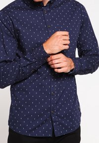 Marineblauwe button-up shirt met een klein wit bladerenpatroon, lange mouwen en een spread kraag. Voorzien van een borstzak en knoopmanchetten.