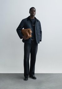 Μπουφάν σκούρου μπλε, μαύρο μπλουζάκι, παντελόνι από σκούρο τζιν και μαύρα παπούτσια. Κρατάει καφέ δερμάτινη τσάντα με πτυσσόμενο σχέδιο. Ομαλές υφές σε όλο το σύνολο.