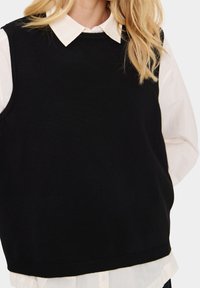 Kvinna som bär en svart ärmlös tröja över en vit skjorta med långa ärmar och blont vågigt hår.