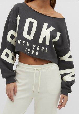 Frau trägt ein dunkelgraues, kurz geschnittenes Sweatshirt mit der Aufschrift "BROOKLYN NEW YORK 1986" und weiße Jogginghosen mit Kordelzug.