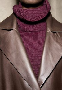 Cappotto in pelle marrone con un colletto ampio sovrapposto a un dolcevita a coste bordeaux. I contrasti di texture tra materiali lisci e lavorati a maglia.