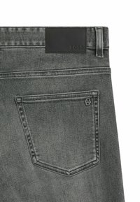 Spatele blugilor gri din denim cu un buzunar cusut, logo mic din metal pe buzunar, buclă pentru curea și eticheta de marcă rectangulară neagră deasupra buzunarului.