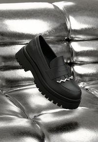 Black leather loafer with a chunky, textured sole. Features a decorative silver zigzag accent on the front. Positioned against a shiny silver background.