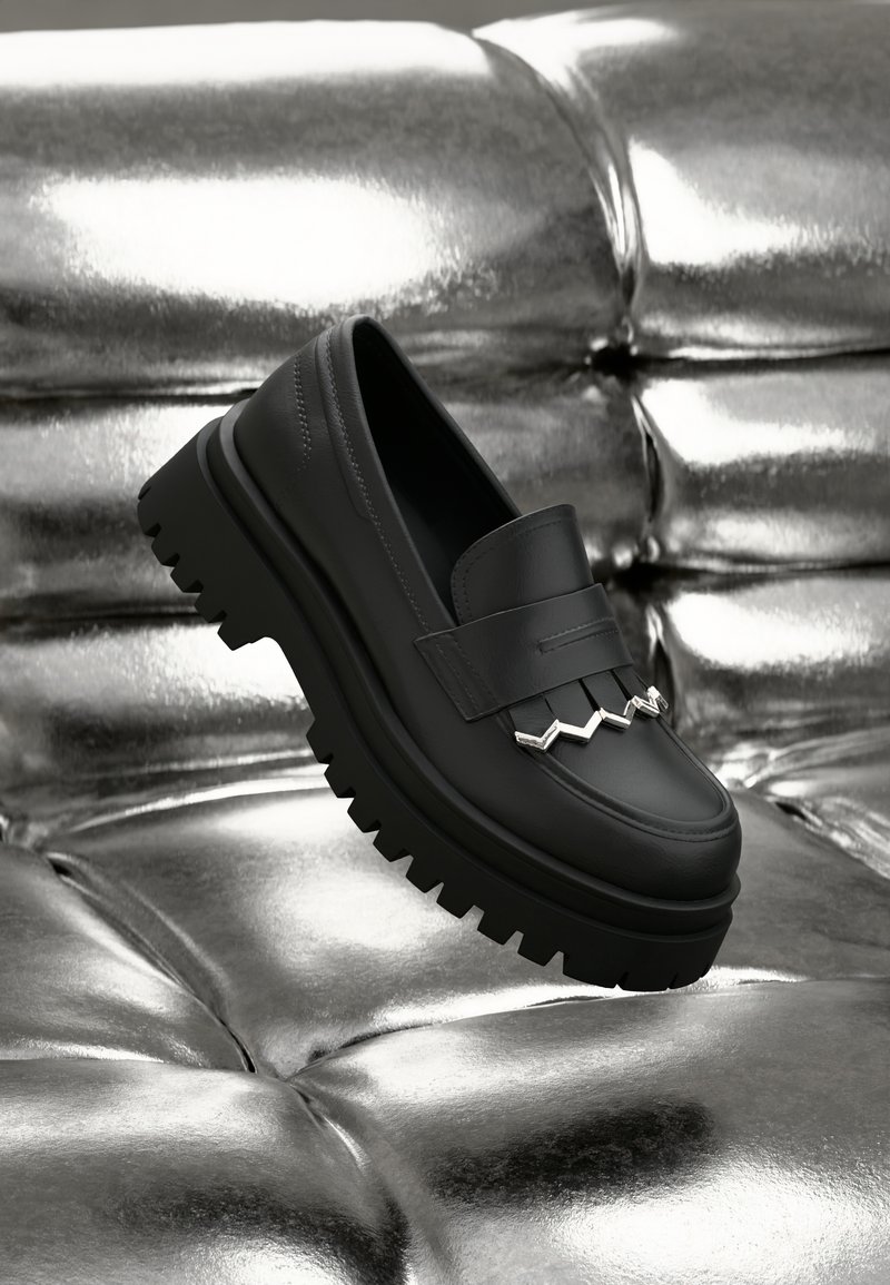 Black leather loafer with a chunky, textured sole. Features a decorative silver zigzag accent on the front. Positioned against a shiny silver background.