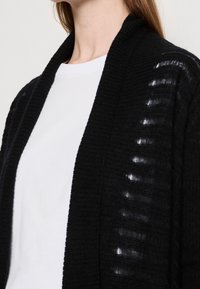 Svart cardigan med en texturerad stickad design, med en avslappnad passform och öppen front. Den har horisontella utskärningar längs ärmarna.