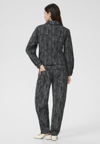 Femme debout avec la main dans la poche arrière, portant une veste texturée gris foncé et un pantalon assorti, des chaussures à talons carrés blanches, tournant le dos.