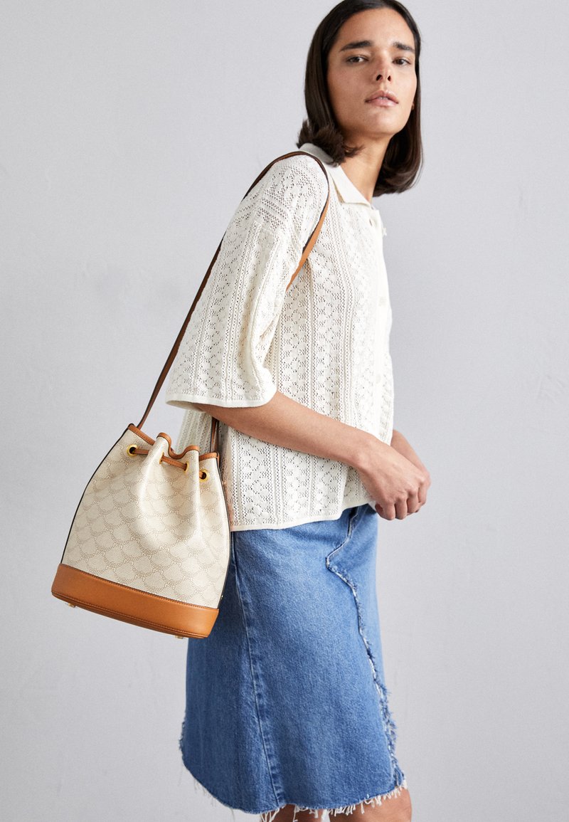 Canvas bucket bag in beige with a patterned exterior, tan leather base, adjustable drawstring, and contrasting handles. Worn across the body.