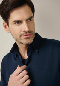 Granatowa koszulka polo o miękkiej fakturze, z kołnierzykiem zapinanym na guziki, eleganckim krojem i delikatnym połyskiem. Model lekko trzyma kołnierzyk.