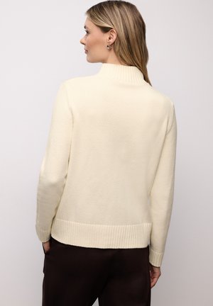 Cremefarbener Pullover mit hohem Rippkragen, geripptem Saum und langen Ärmeln. Weiche Textur sichtbar, kombiniert mit dunklen Hosen.