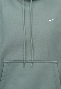 Sivasto-zelená mikina s klokaním vreckom, šnúrkami na stiahnutie a malým bielym logom Nike na hrudi. Mäkká, textúrovaná tkanina.