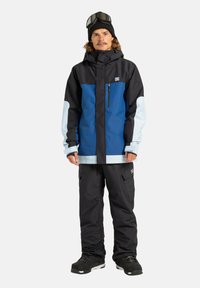 Niebieska i czarna kurtka śniegowa z jasnoniebieskimi akcentami, zamykanymi kieszeniami i regulowanymi mankietami, w połączeniu z czarnymi spodniami śniegowymi i butami.