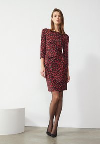 Rot- und schwarz gemustertes Kleid mit Dreiviertelärmeln, taillierter Silhouette und gerafften Details an der Taille. Getragen mit transparenten schwarzen Strumpfhosen.