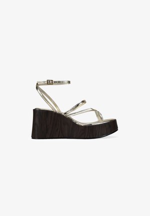 Sandalia de cuña con plataforma de madera oscura y finas tiras metálicas doradas, incluyendo una correa ajustable en el tobillo con una pequeña hebilla.