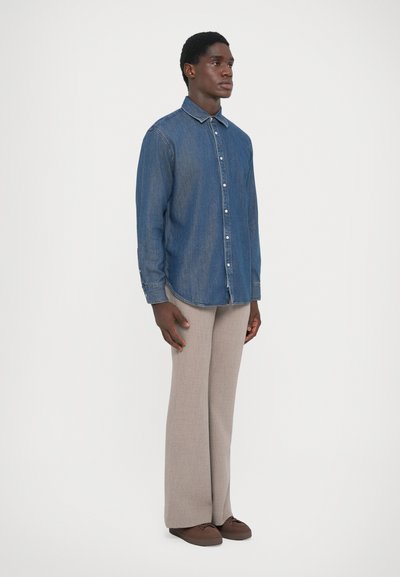 Chemise en denim bleu à manches longues avec fermeture à boutons et col, associée à un pantalon beige et des chaussures brunes. Design simple et classique.