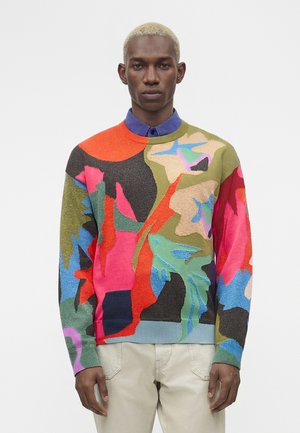 Mężczyzna z krótkimi blond włosami, ubrany w kolorowy sweter z abstrakcyjnym wzorem na niebieskiej koszuli z kołnierzykiem i jasnobeżowych spodniach, stojący neutralnie.