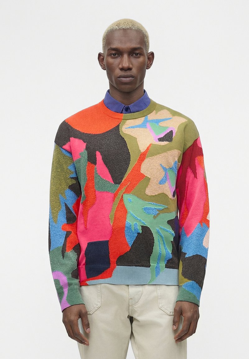 Άνδρας με κοντά ξανθά μαλλιά που φοράει πολύχρωμο πουλόβερ με αφηρημένο σχέδιο πάνω από ένα μπλε πουκάμισο με γιακά και ανοιχτό μπεζ παντελόνι, στέκεται ουδέτερα.