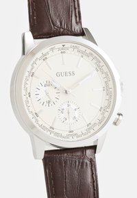 Montre argentée avec un cadran crème, un bracelet en cuir marron et plusieurs sous-cadrans. Comprend des index horaires et les noms de villes du monde autour de la lunette.