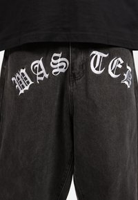 Pantalones de mezclilla negros con gran texto bordado en blanco que dice "WASTED" en la parte frontal. Costuras visibles y un ajuste relajado.