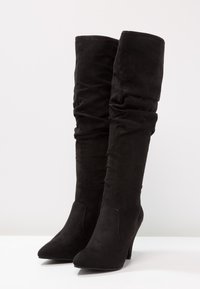 Botas negras de ante hasta la rodilla, con un diseño holgado, punteras afiladas y un tacón alto para altura y estilo.
