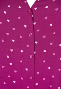 Fuchsiablus med V-ringning, prydd med ett mönster av små rosa och vita hjärtan. Fem rosa knappar på framsidan. Slät textur.