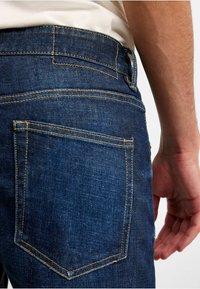 Mörkblå denimjeans med kontrasterande gult stygn, en bakficka med rundad form och en standard midjebandsdesign.