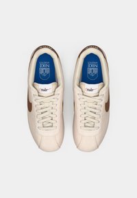 Par bežnih Nike športnih copat z rjavim swooshom, belimi vezalkami in modrimi notranjostmi, označenimi z "Nike Athletics", prikazani od zgoraj na belem ozadju.