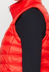 Červená prošívaná vesta s lesklou syntetickou látkou, bez rukávů, s vysokým límcem, pod ní černý žebrovaný top.