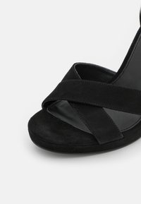 Gros plan d'une sandale noire en suédine à bout ouvert avec deux brides croisées et une semelle rembourrée sur fond blanc.